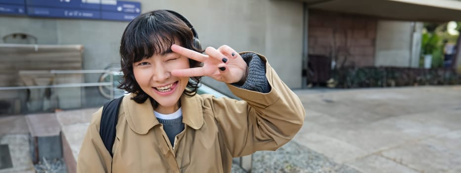 Head portrait of korean girl standing on street, showing peace sign, wearing headphones, listening music and smiling.