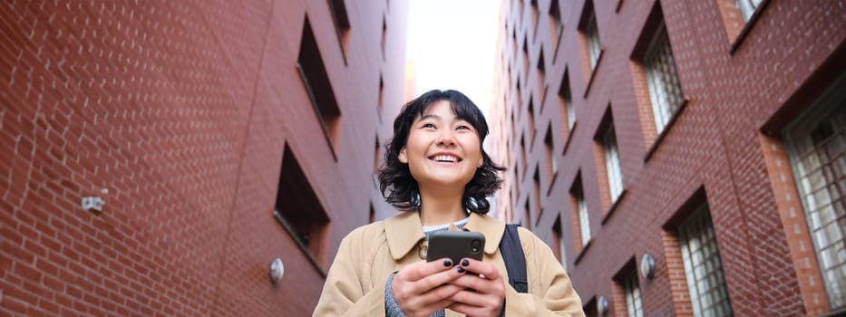 Lower angle view of brunette korean girl, listening music in headphones, walking along street and looking at smartphone, reading message on mobile phone.