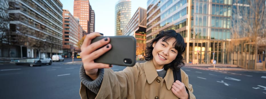 Portrait of young asian woman taking selfie in front of building in city centre, tourist takes photos while sightseeing, smiling at smartphone camera.