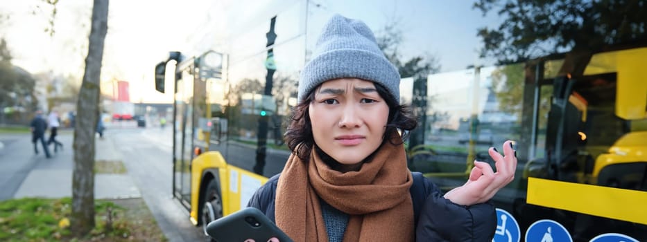 Portrait of confused asian girl, standing on bus stop, holding mobile phone, looking shocked and upset, disappointed by public transport delay.