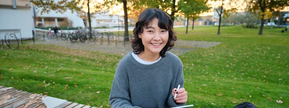 Portrait of young smiling korean girl, graphic designer, artist drawing on digital tablet with a pen tool, sitting in park on fresh air and scatching, taking notes.