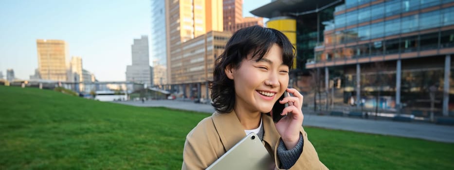 Stylish smiling girl, university student in trench coat, holds tablet, talks on mobile phone, has conversation over telephone and looks relaxed, stands on street.