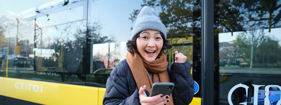 Enthusiastic girl rejoice, reads message on mobile phone and celebrates, stands near her bus on public transport stop and looks excited, posing in warm winter clothes.