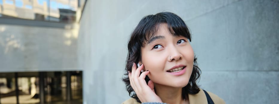 Close up portrait of smiling korean girl in headphones, listens music in wireless earphones, enjoys travelling around town with her favorite songs playlist.