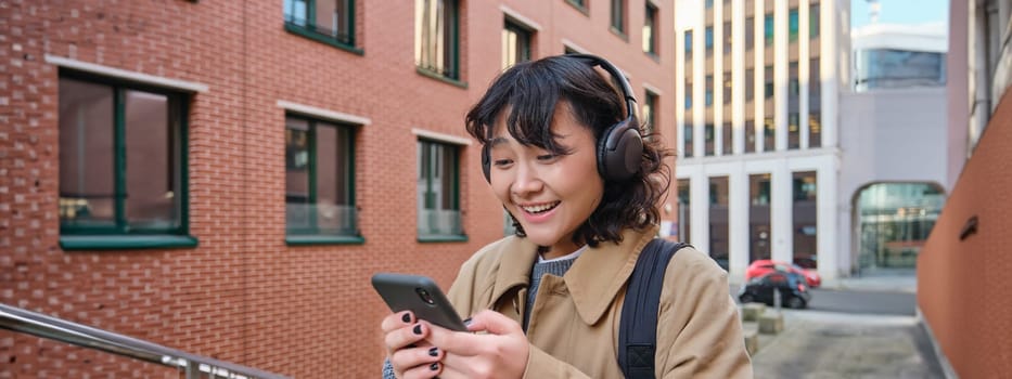 Happy korean girl looks at mobile phone and listens music in headphones, stands on street in city centre, reads text message on smartphone.