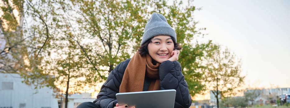 Portrait of asian girl relaxing in park, watching videos or reading on digital tablet, sitting on bench in hat and scarf on chilly day, enjoing outdoors.