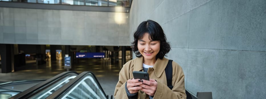 Smiling korean girl, student with smartphone goes up an escalator, reads mobile phone text message.