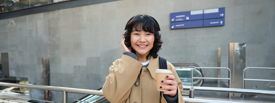 Beautiful smiling Korean girl, drinks takeaway coffee, listens music in headphones, wears trench and backpack, stands on train station, travelling.