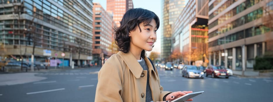 Woman walking on street using digital tablet, holding gadget in hands and looking away, standing in city centre.