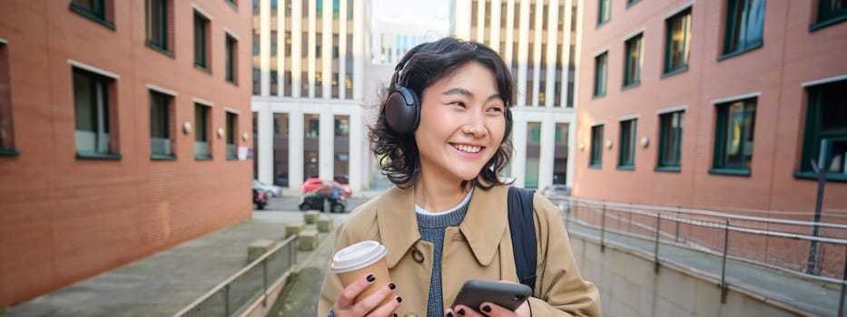 Young woman, tourist in headphones looks at smartphone, drinks coffee to go, checks mobile phone app, listens music and travels. Copy space