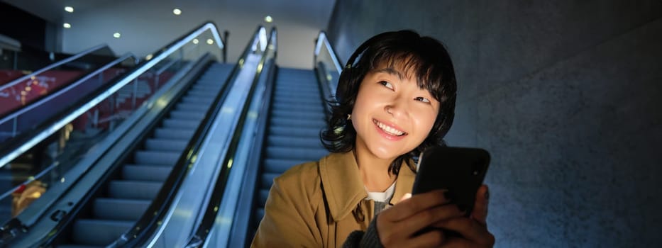 Smiling korean girl going down escalator in dark, holding mobile phone, using smartphone app, listening music, commuting in city.