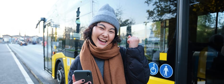 Enthusiastic girl rejoice, reads message on mobile phone and celebrates, stands near her bus on public transport stop and looks excited, posing in warm winter clothes.