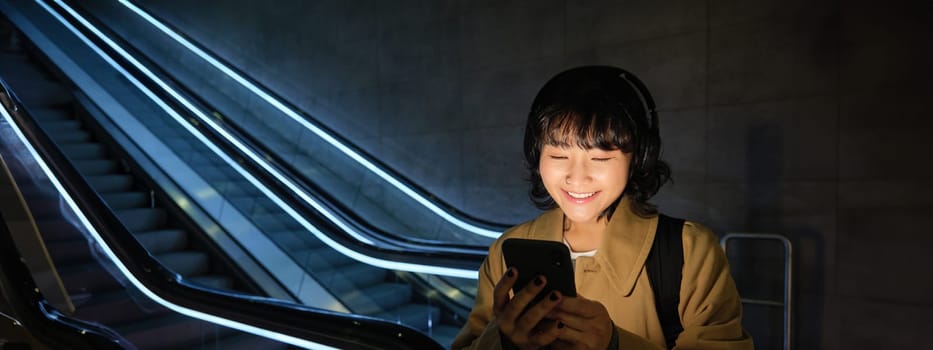 Happy asian woman in subway, standing near escalator, holding smartphone and listening music in headphones, smiling pleased.