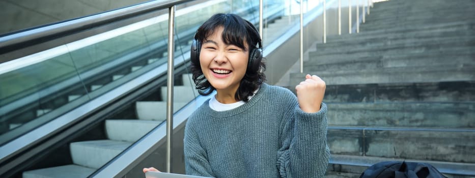 Cheerful korean girl in headphones, sits on stairs with laptop, celebrates, say yes, triumphs, receives good news, success on working project, works remotely on street.