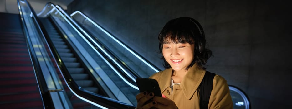 Portrait of young smiling asian girl, stands near escalator and waits for someone, reads message, checks map on smartphone, listens music in headphones.