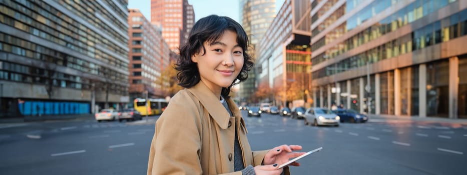 Woman walking on street using digital tablet, holding gadget in hands and looking away, standing in city centre.