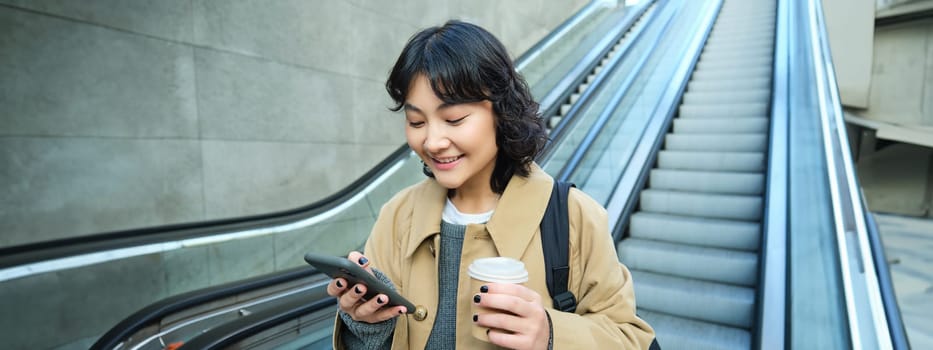 Young stylish girl with cup of coffee, drinks cappuccino to go, goes down escalator and looks at mobile phone, chats, reads message on smartphone.