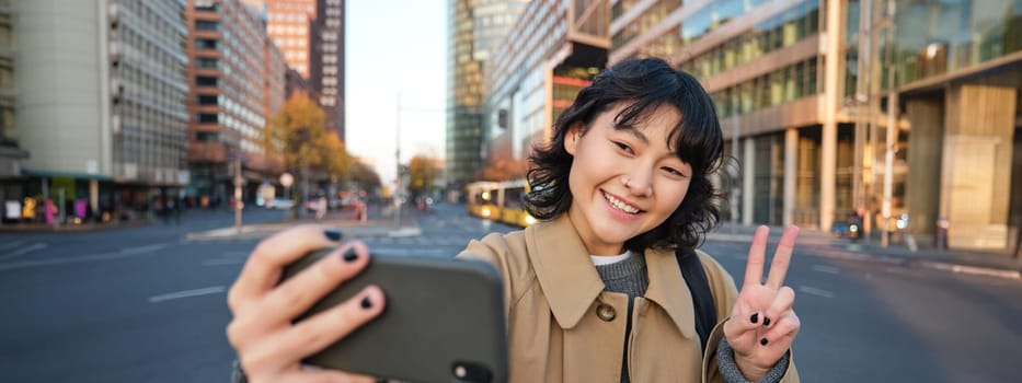 Happy asian girl, tourist takes photo in city centre, shows peace sign at smartphone camera, makes selfie during sightseeing with smiling joyful face.