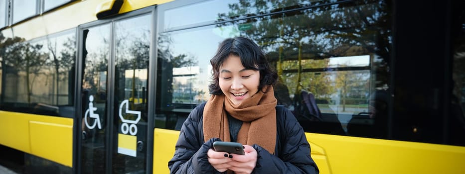 Beautiful korean girl, student on bus stop, looking at her smartphone, checking timetable, reading text message, wearing winter clothes.