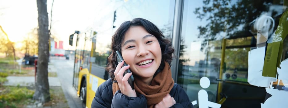 Cellular technology and people concept. Stylish asian girl talks on mobile phone, makes a telephone call, stands near bus stop and has conversation. Copy space