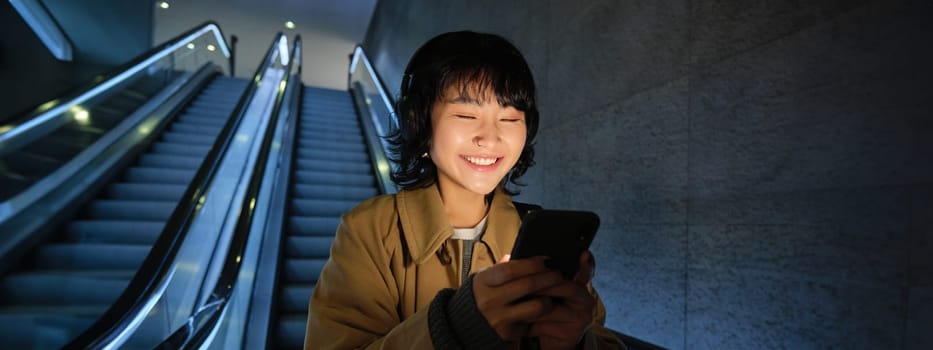 Smiling korean girl going down escalator in dark, holding mobile phone, using smartphone app, listening music, commuting in city.