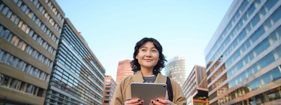 Portrait of happy korean girl smiles, walks in city with digital tablet and backpack, uses her gadget to get around.