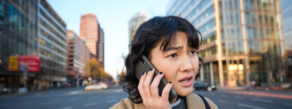 Close up portrait of concerned asian woman, talks on mobile phone and hears bad news, looks worried, feels frustrated by telephone conversation, stands on street.