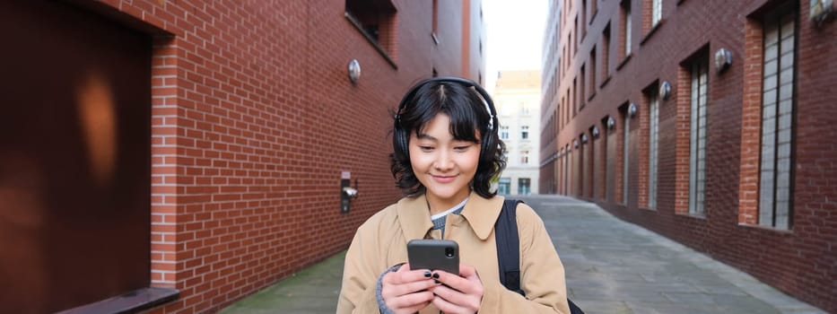 Young asian woman listens music in wireless earphones, uses smartphone on street, walks around city on sunny day, smiling happily.