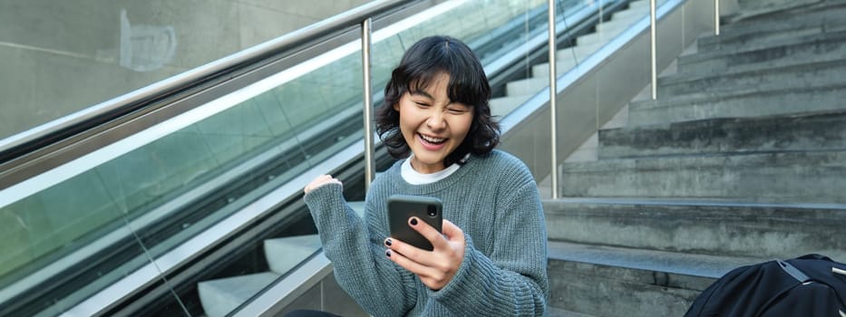 Portrait of brunette young woman, student sits on stairs in public place, looks at smartphone, reads text message and smiles.