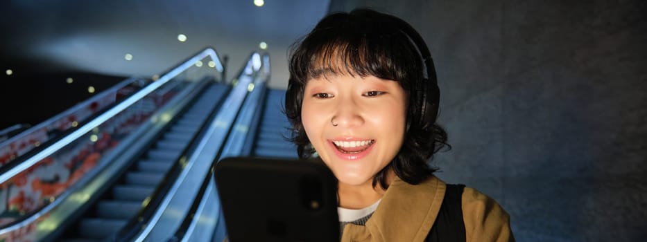 Close up of happy young asian girl, looks at her smartphone with surprised, excited face expression, reading good news on phone, standing on escalator in city.