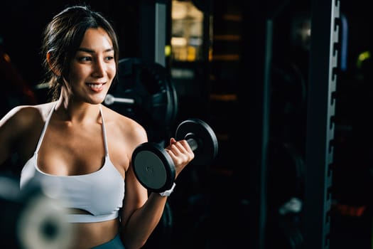 A smiling Asian woman in sportswear, lifting weights and holding a dumbbell, showcases her athletic biceps and strong body for a fitness portrait in the gym