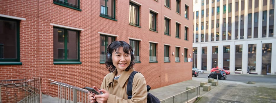 Young happy woman in trench coat, listens music in headphones, holds smartphone, uses mobile application to find route in new city, stands on street with backpack.