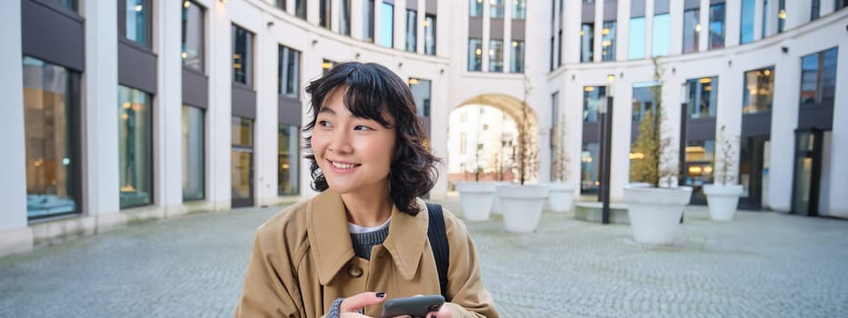 Stylish korean girl in headphones, listens music and uses mobile phone, stands in city centre, waits for someone on street and writes text message on her smartphone.