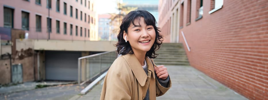 Young asian woman listens music in wireless earphones, uses smartphone on street, walks around city on sunny day, smiling happily.