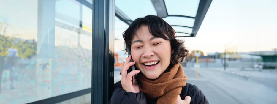 Close up of asian girl on bus stop, laughs over phone conversation, talks on telephone while waits for public transport, wears winter clothes.