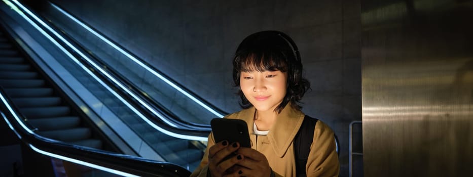 Portrait of young smiling asian girl, stands near escalator and waits for someone, reads message, checks map on smartphone, listens music in headphones.