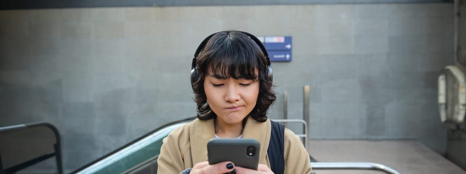 Portrait of asian girl in headphones, looks complicated at smartphone screen, puzzled by text message or notification, stands on street and shrugs.