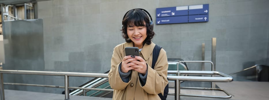 Smiling asian girl in headphones, stands on train station, looks at mobile phone screen and looks pleased, listens music and drinks coffee.