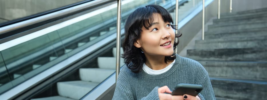 Stylish young girl student, sits on stairs with smartphone and backpack, laughs and smiles, texts message, uses social media.