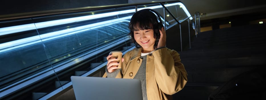 Beautiful young girl student, korean woman sits on staircase, watches video on laptop in headphones, drinks takeaway coffee, works in public place on remote.