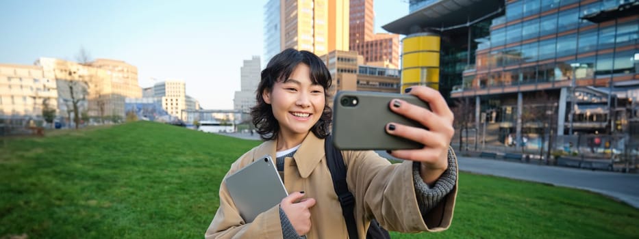 Happy university student, korean girl takes selfie with her papers and digital tablet, holds smartphone and poses near university campus.