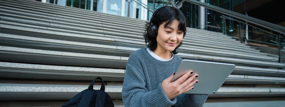 Young asian girl, student in headphones, works on remote, digital artist drawing on tablet with graphic pen, listening music in headphones and sitting on street staircase.