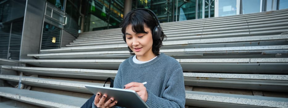 Young smiling digital artists works on her project using tablet and graphic pen, listens music in headphones and sits on stairs in public space, works on project.