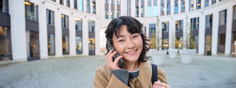 Portrait of smiling asian girl talks on mobile phone, speaks with friend on smartphone, walks in city centre, stands on street and laughs.