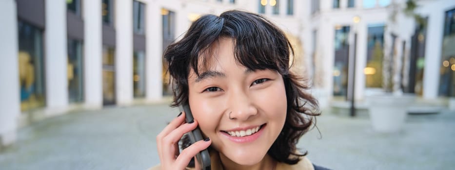 Portrait of smiling asian girl talks on mobile phone, speaks with friend on smartphone, walks in city centre, stands on street and laughs.