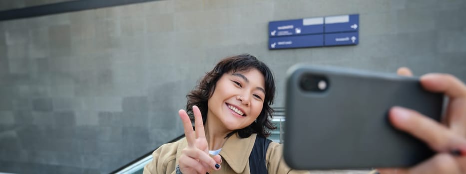 Portrait of stylish korean girl, student takes selfie on street, holds smartphone and poses on its camera, makes video on tourist trip, smiles with joy.