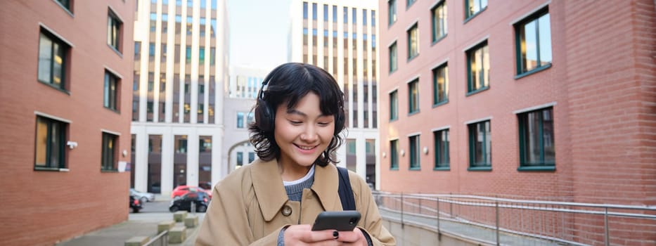 Portrait of cute girl tourist, korean woman in headphones, looks at mobile phone, uses smartphone app, map or text messages, listens music in headphones.