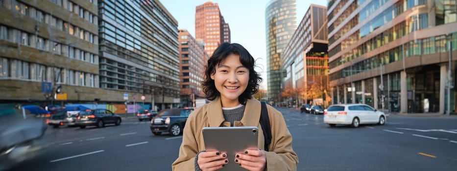 Portrait of asian girl student, stands in city centre with cars on busy street, holds digital tablet and smiles at camera.