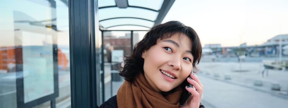 Close up of cute Korean woman, making a phone call, talking and smiling on telephone, standing in winter jacket on bus stop, waiting for her transport to arrive.