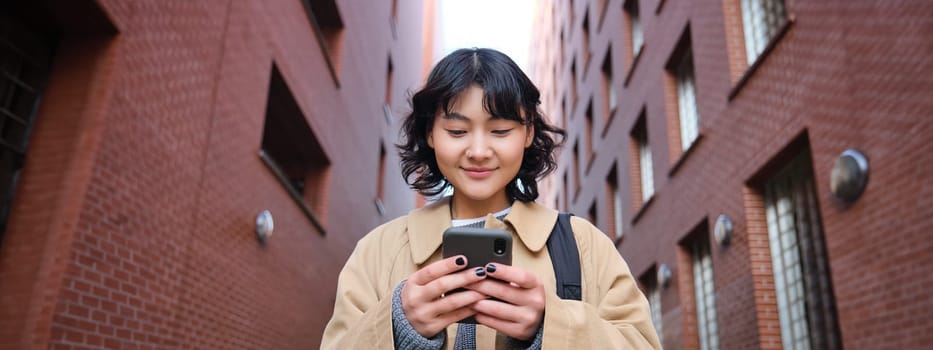 Lower angle view of brunette korean girl, listening music in headphones, walking along street and looking at smartphone, reading message on mobile phone.
